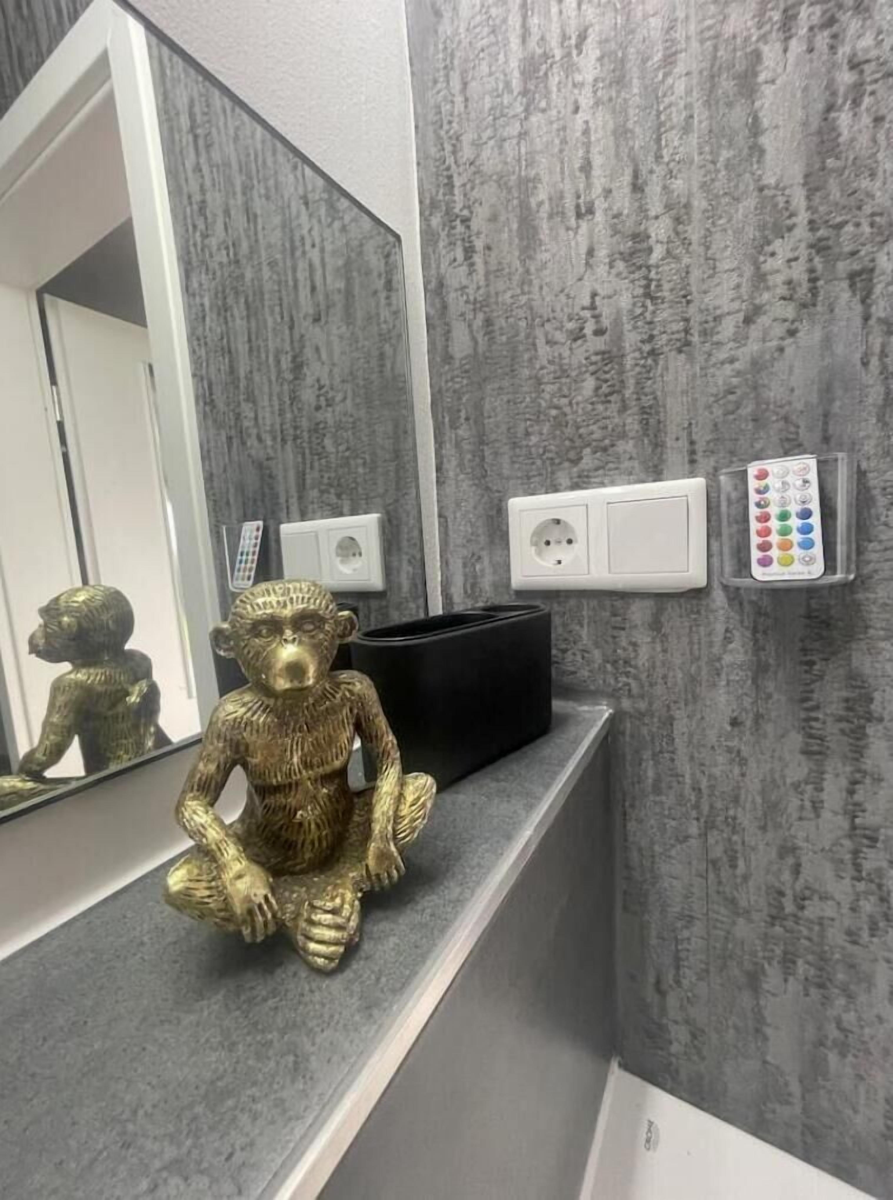Ein Badezimmer mit einer grauen Wand mit Struktur, einem Spiegel, einer goldenen Statue, einer schwarzen Tasche und einer wandmontierten Steckdose mit Fernbedienung.