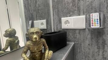 Ein Badezimmer mit einer grauen Wand mit Struktur, einem Spiegel, einer goldenen Statue, einer schwarzen Tasche und einer wandmontierten Steckdose mit Fernbedienung.