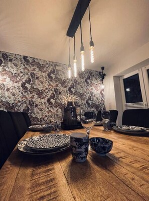 Un comedor con mesa de madera, papel tapiz estampado, sillas negras y lámparas colgantes.