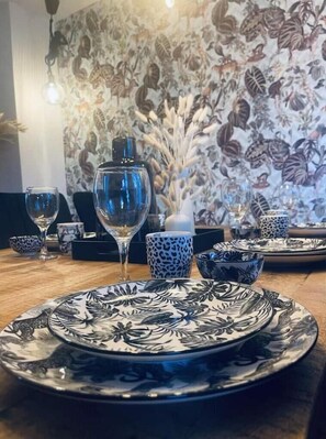 Une table à manger dressée avec des assiettes à motifs, un verre transparent et un vase contenant des fleurs séchées.