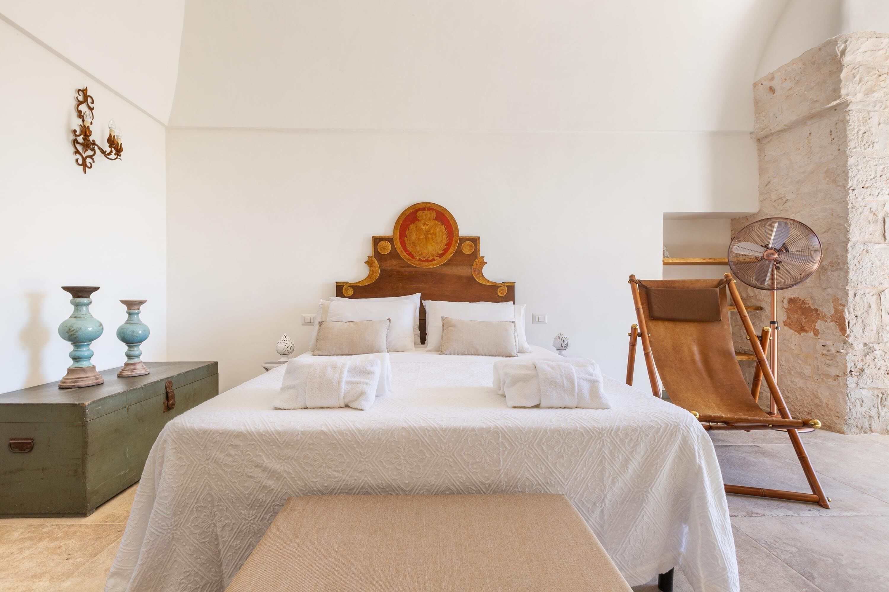 Quarto bem arrumado com uma cama grande, cabeceira de madeira, uma mesinha com objetos decorativos e uma cadeira dobrável.