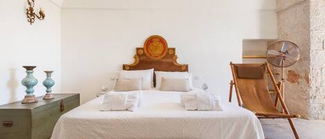 Quarto bem arrumado com uma cama grande, cabeceira de madeira, uma mesinha com objetos decorativos e uma cadeira dobrável.