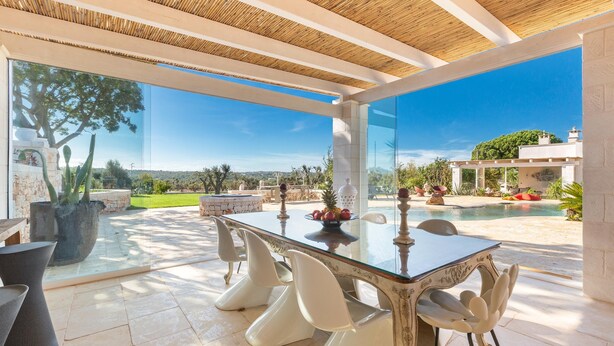 An outdoor dining area with a glass table and white chairs, a pool, and a view of the landscape. - 3666 Ca' Della Roccia by Perle di Puglia (Ostuni)