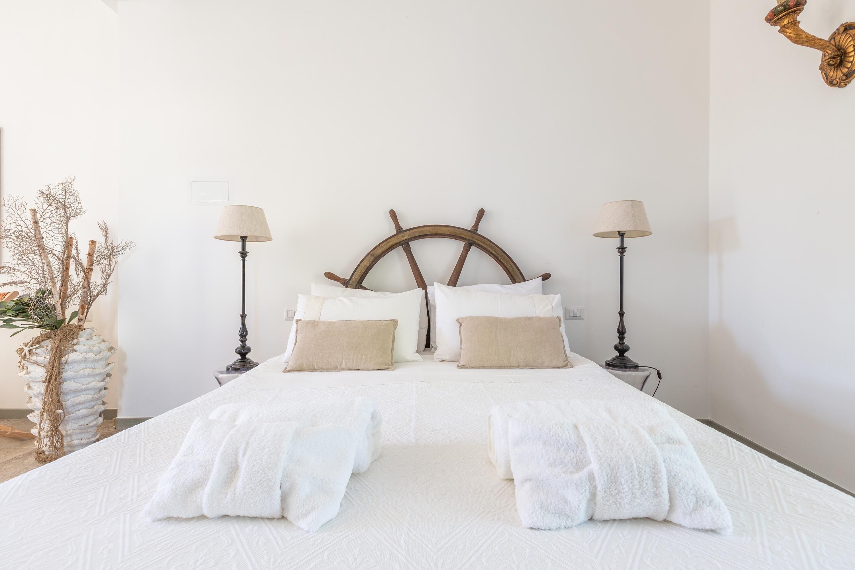 Un lit bien fait, avec du linge de lit blanc et deux oreillers beige, flanqué de deux lampadaires avec des abat-jour beige.