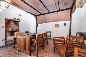 Un área de comedor al aire libre techada, con una mesa y sillas de madera, una parrilla y un estante fijado a la pared con objetos decorativos.