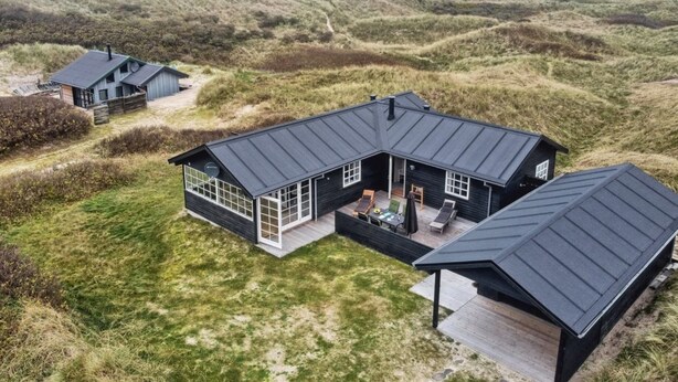 Ett modernt hus med en stor terrass och en inglasad uteplats, omgivet av sanddyner och havet.