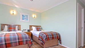 A room with two beds, each with a plaid blanket, a nightstand, a framed picture on the wall, and a door on the right. - Tingle Creek Hotel (Kyle)