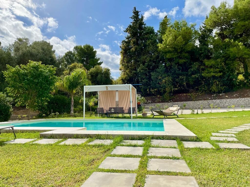 A swimming pool with a covered seating area, surrounded by a stone pathway and lush greenery.