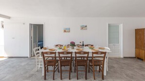 Une salle à manger avec une table blanche dressée pour huit personnes, entourée de chaises en bois. Deux petits tableaux ornent le mur, une porte en verre dépoli se trouve à proximité, et un buffet en bois est placé sur le côté droit.