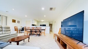 A modern living room with a large flat-screen TV, a sofa, a dining table with chairs, and a kitchen area in the background. - 6 Bedroom House @ Gungahlin Town Center (Gungahlin)