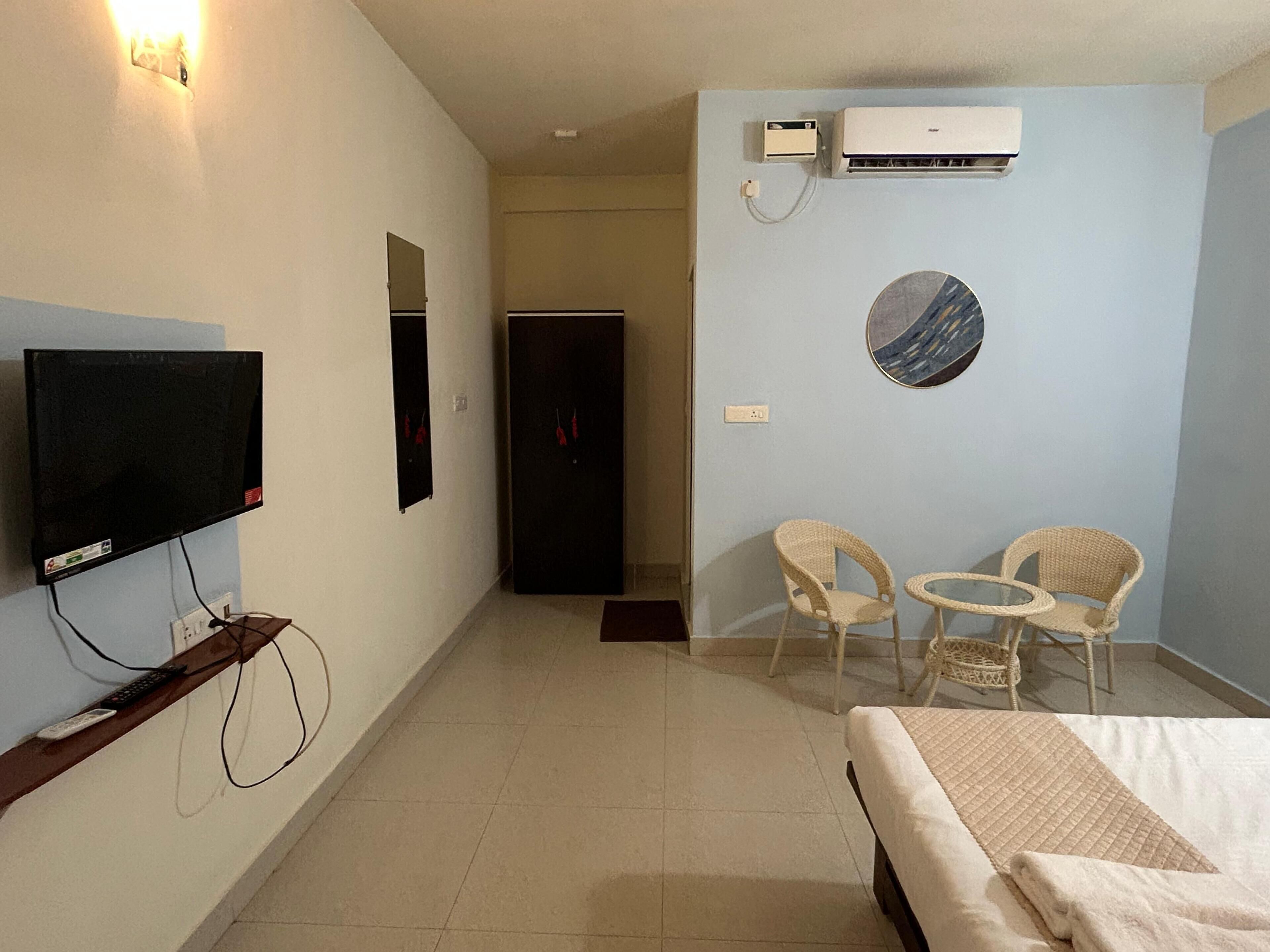 A hotel room with a bed, a television, a wall-mounted air conditioner, a door, and two wicker chairs.