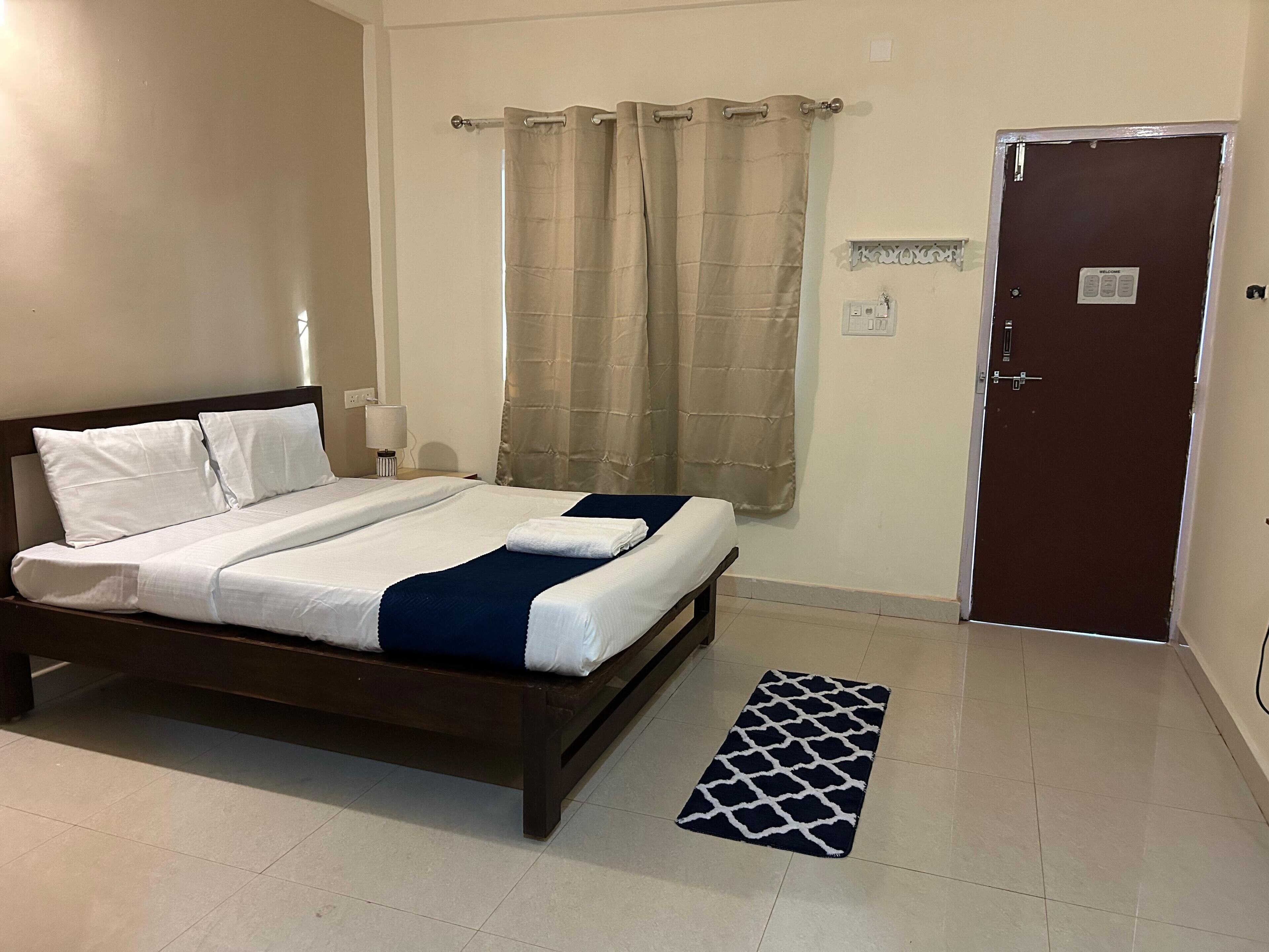A neatly made bed with white and dark blue bedding, a bedside table with a lamp, a window with beige curtains, and a door with a welcome sign.