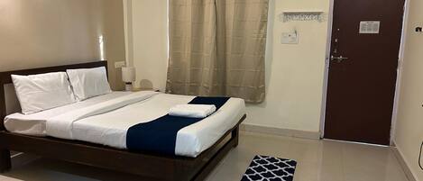 A neatly made bed with white and dark blue bedding, a bedside table with a lamp, a window with beige curtains, and a door with a welcome sign.