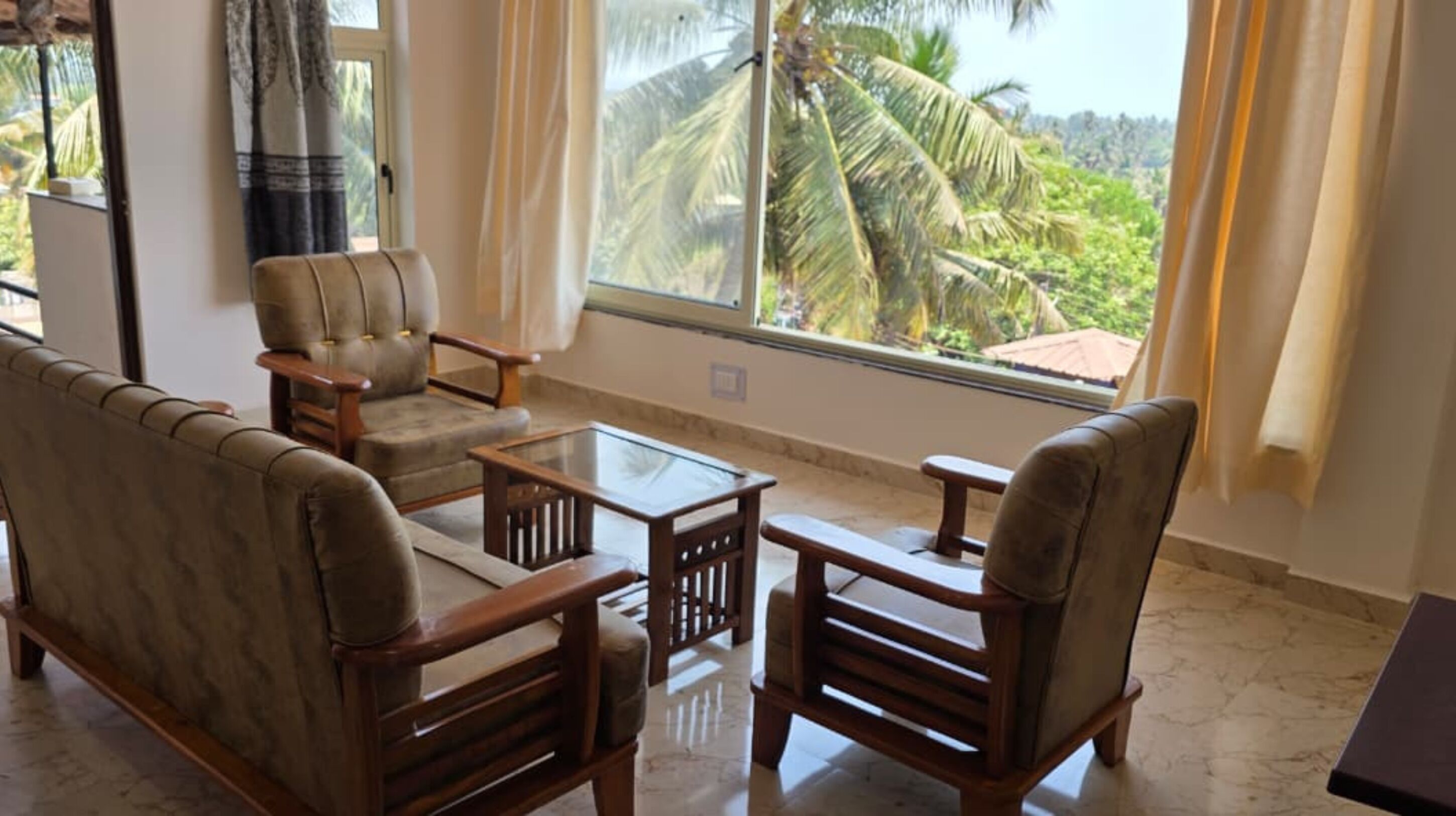 A room with a sofa, two armchairs, a glass coffee table, and a view of palm trees through large windows.