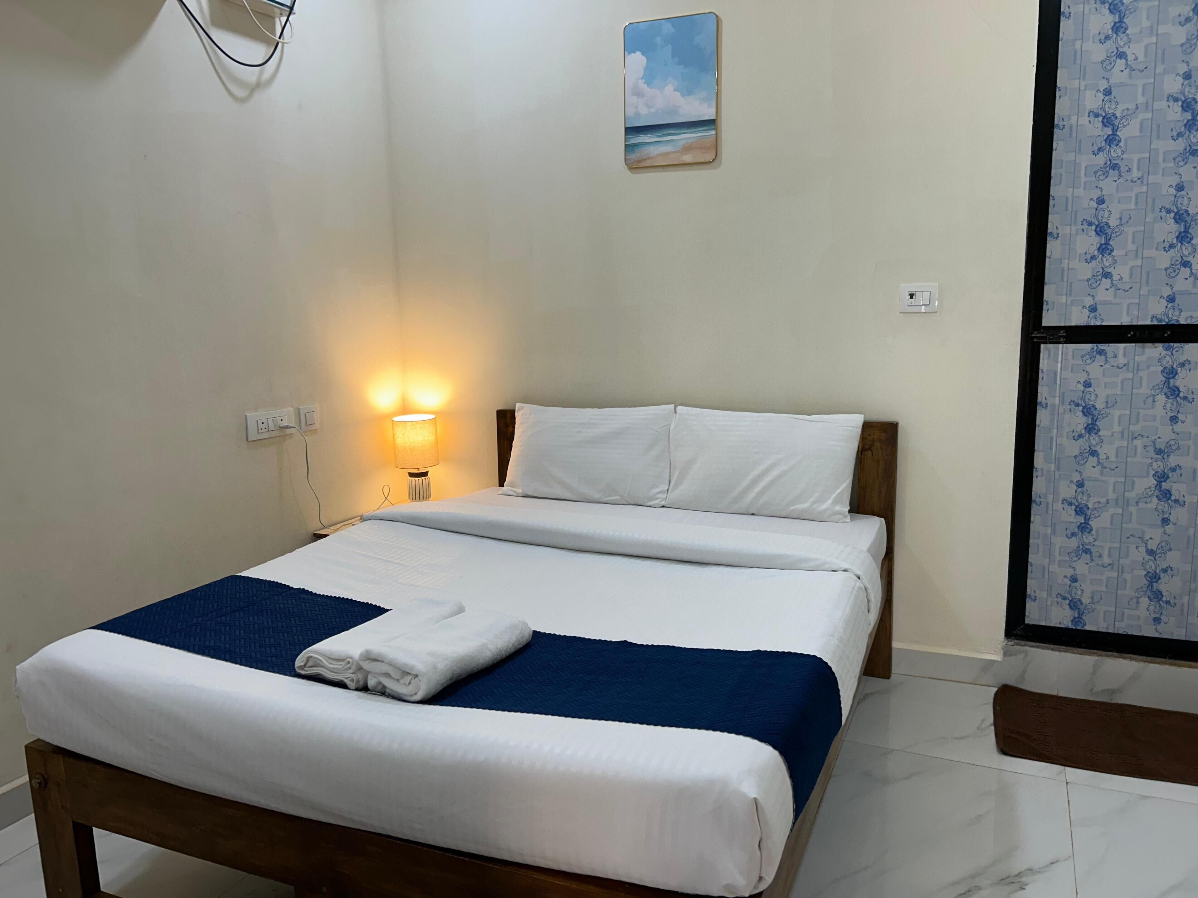 A neatly made bed with white linens and a blue blanket, a bedside lamp, and a wall-mounted painting of a beach scene.