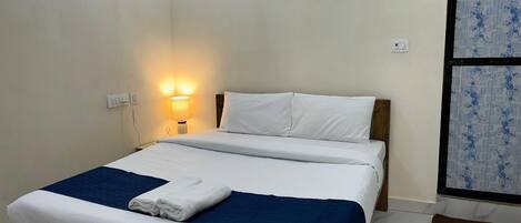 A neatly made bed with white linens and a blue blanket, a bedside lamp, and a wall-mounted painting of a beach scene.