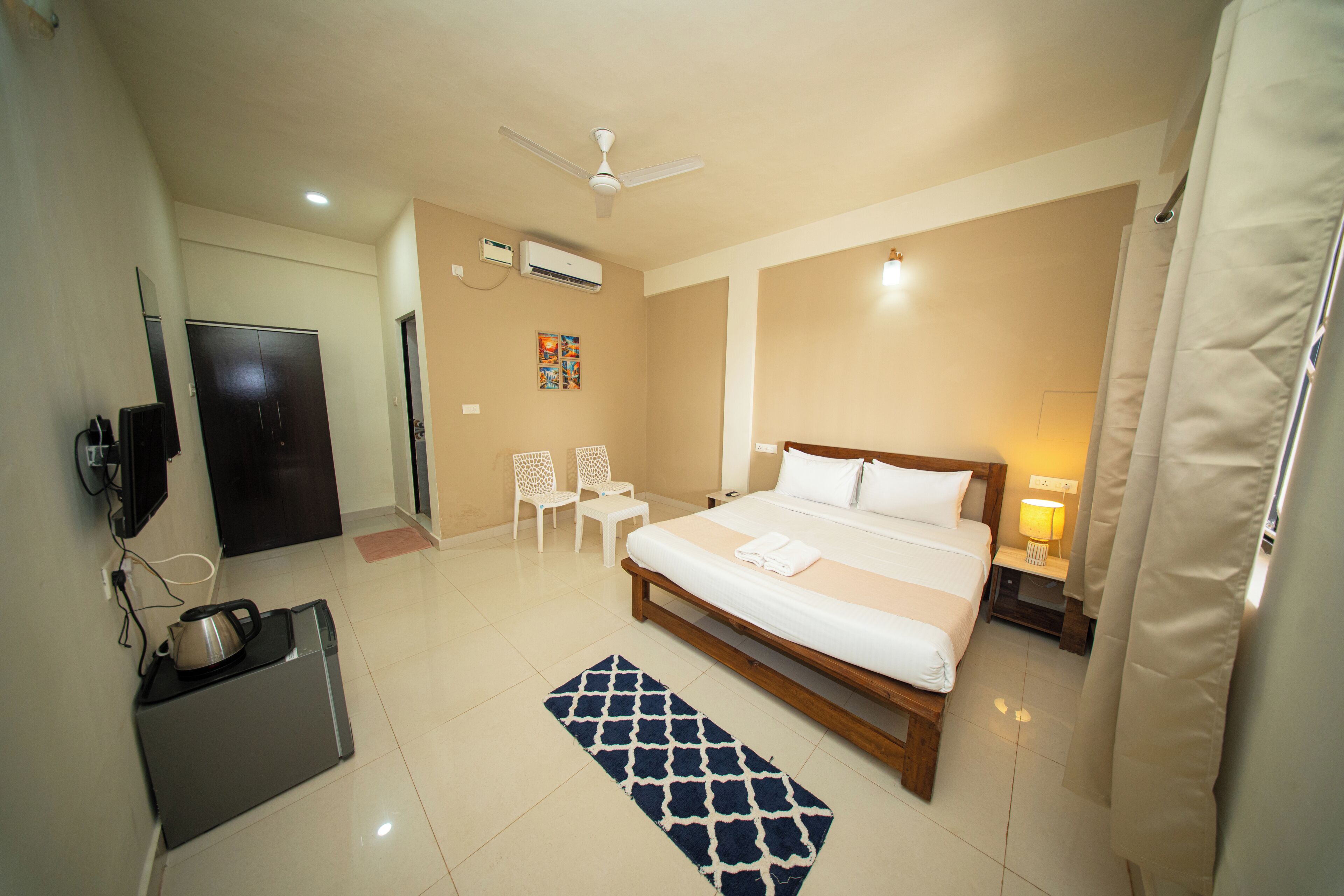 A hotel room with a bed, a desk with a kettle, a TV, a ceiling fan, and an air conditioner.