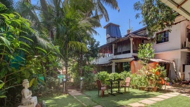 A courtyard with a statue, a table, and chairs, surrounded by lush greenery and a building with a water tank on the roof. - Aqua Wave by YogiTree Hotels (Morjim)