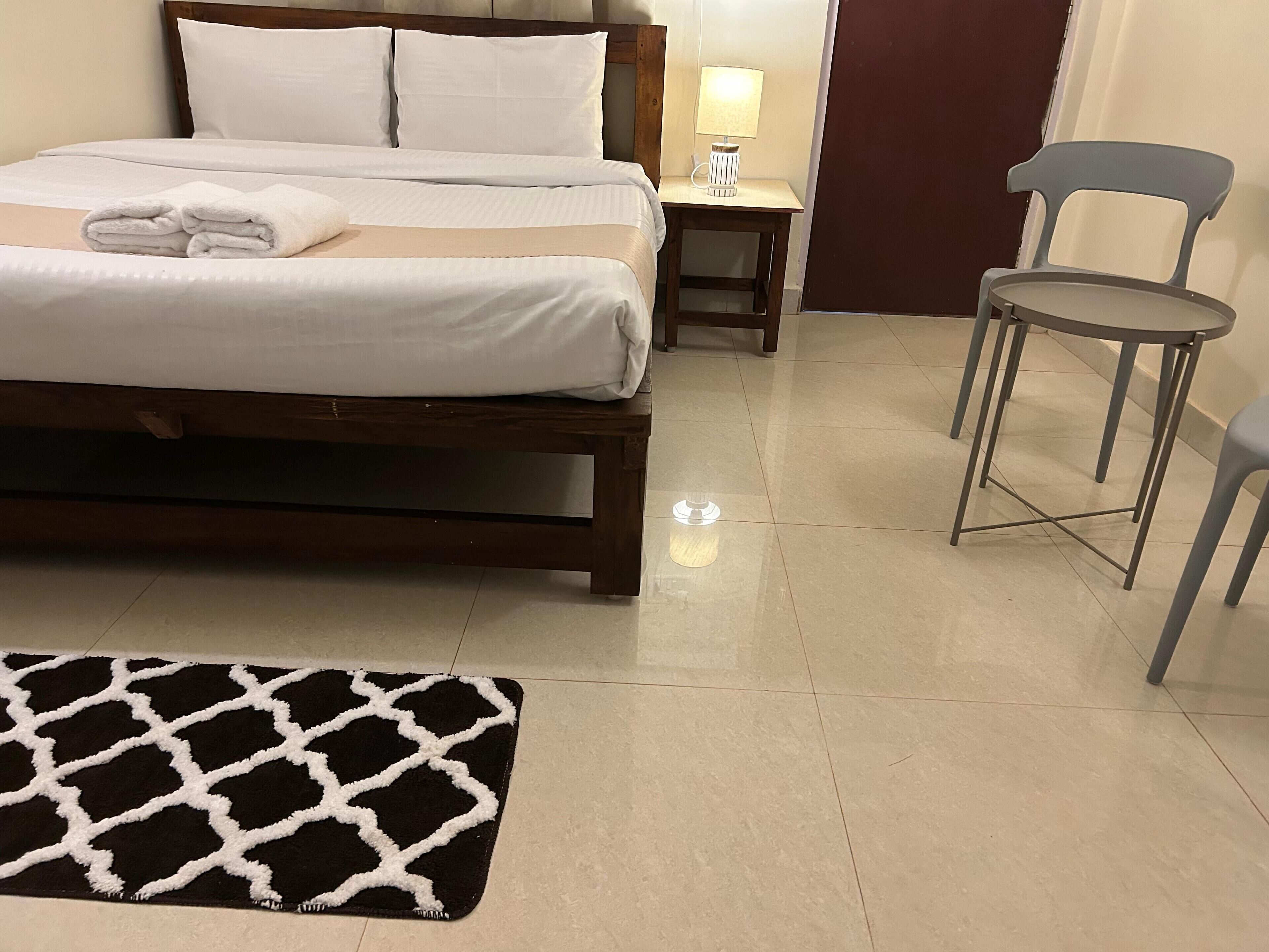 Un lit bien fait, avec du linge de lit blanc, une table de chevet avec une lampe, deux chaises et un tapis noir et blanc.
