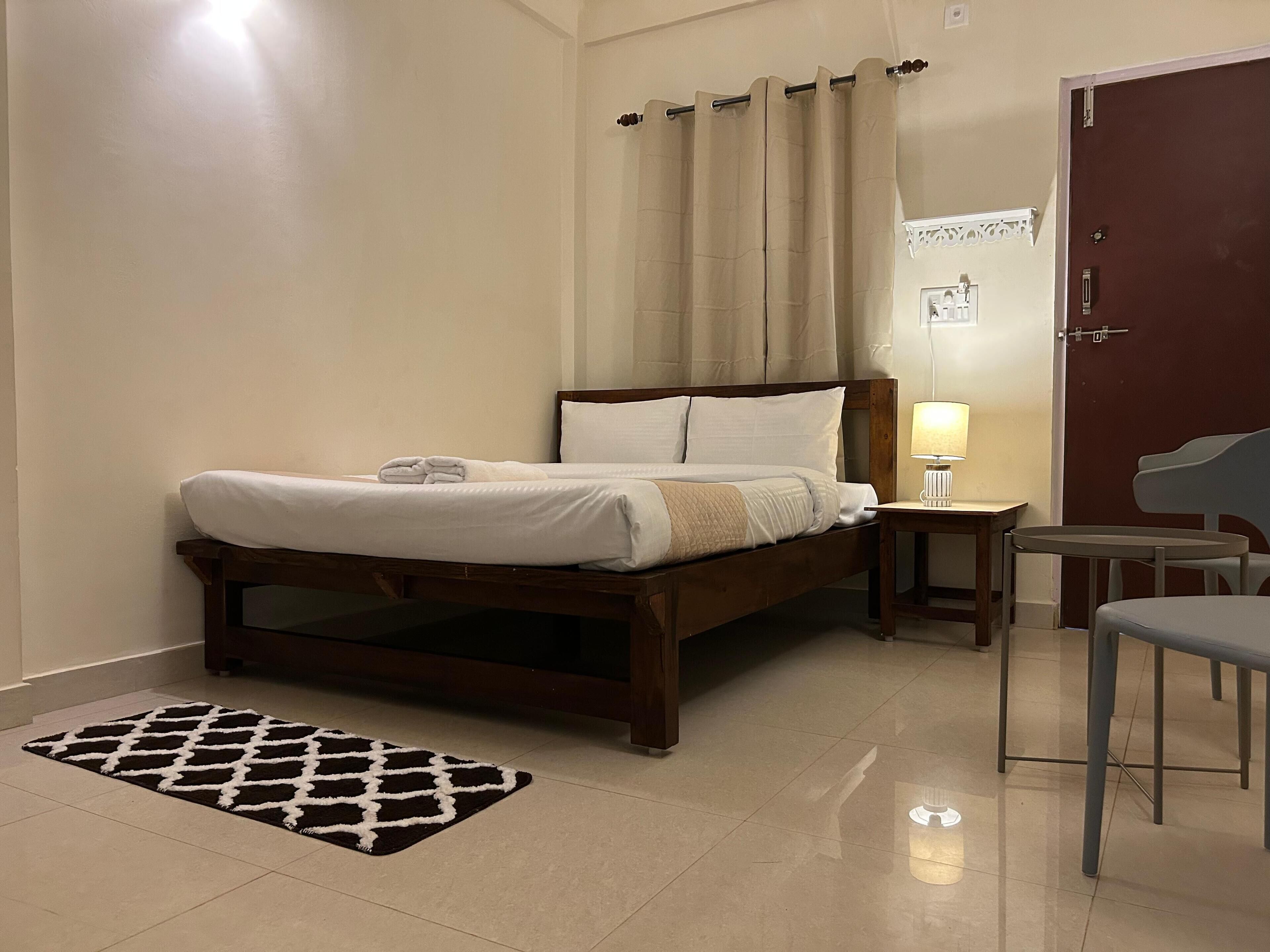 Quarto bem arrumado com cama, mesa de cabeceira, abajur, cadeira e tapete estampado.