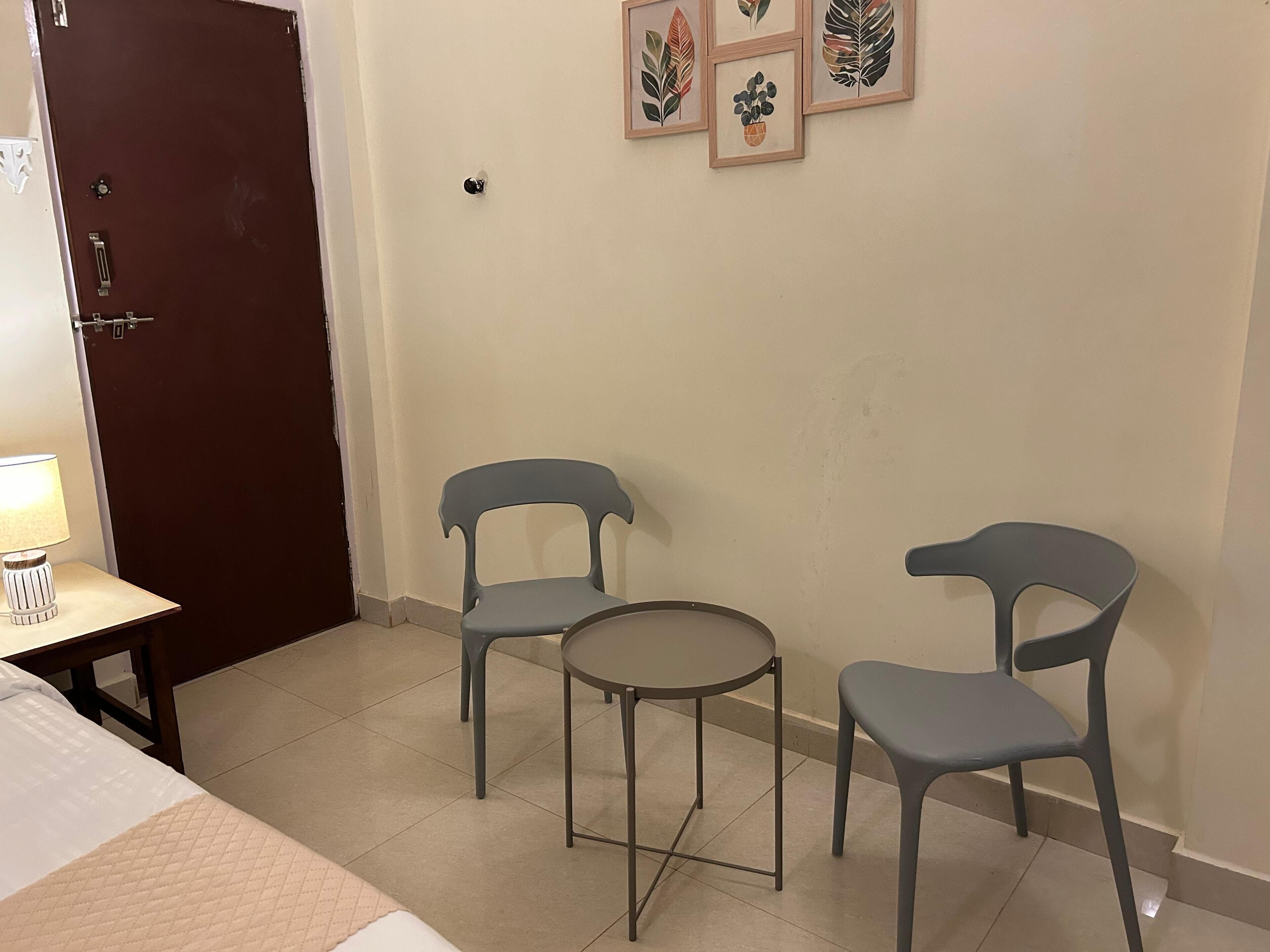 A room with a bed, a small table with a lamp, two chairs, and a door.