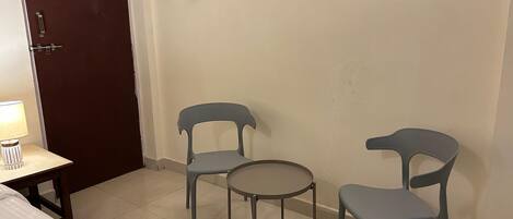 A room with a bed, a small table with a lamp, two chairs, and a door.