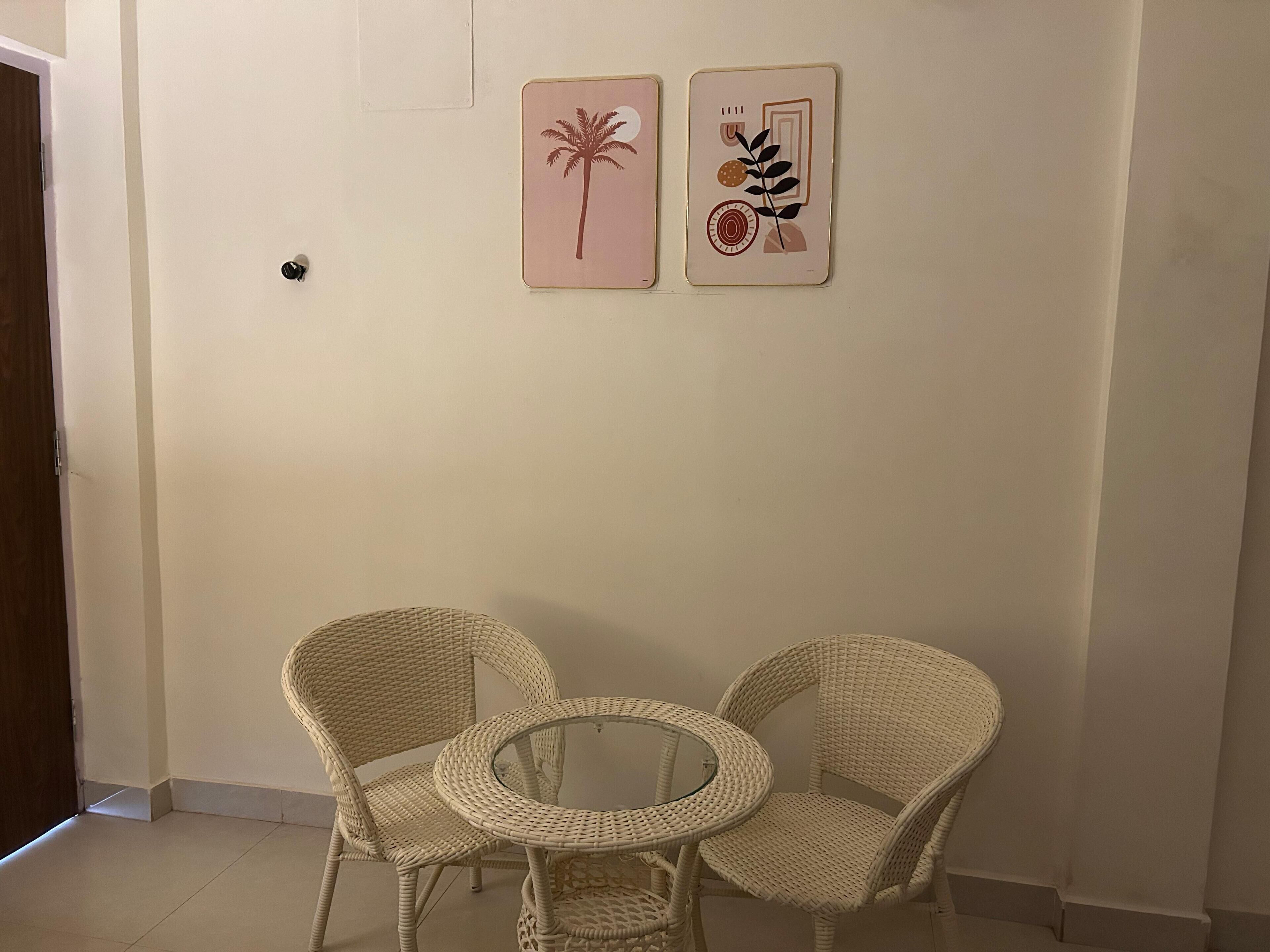 Una habitación con dos sillas de mimbre, una mesita redonda pequeña y una puerta de madera, además de dos cuadros colgados en la pared.