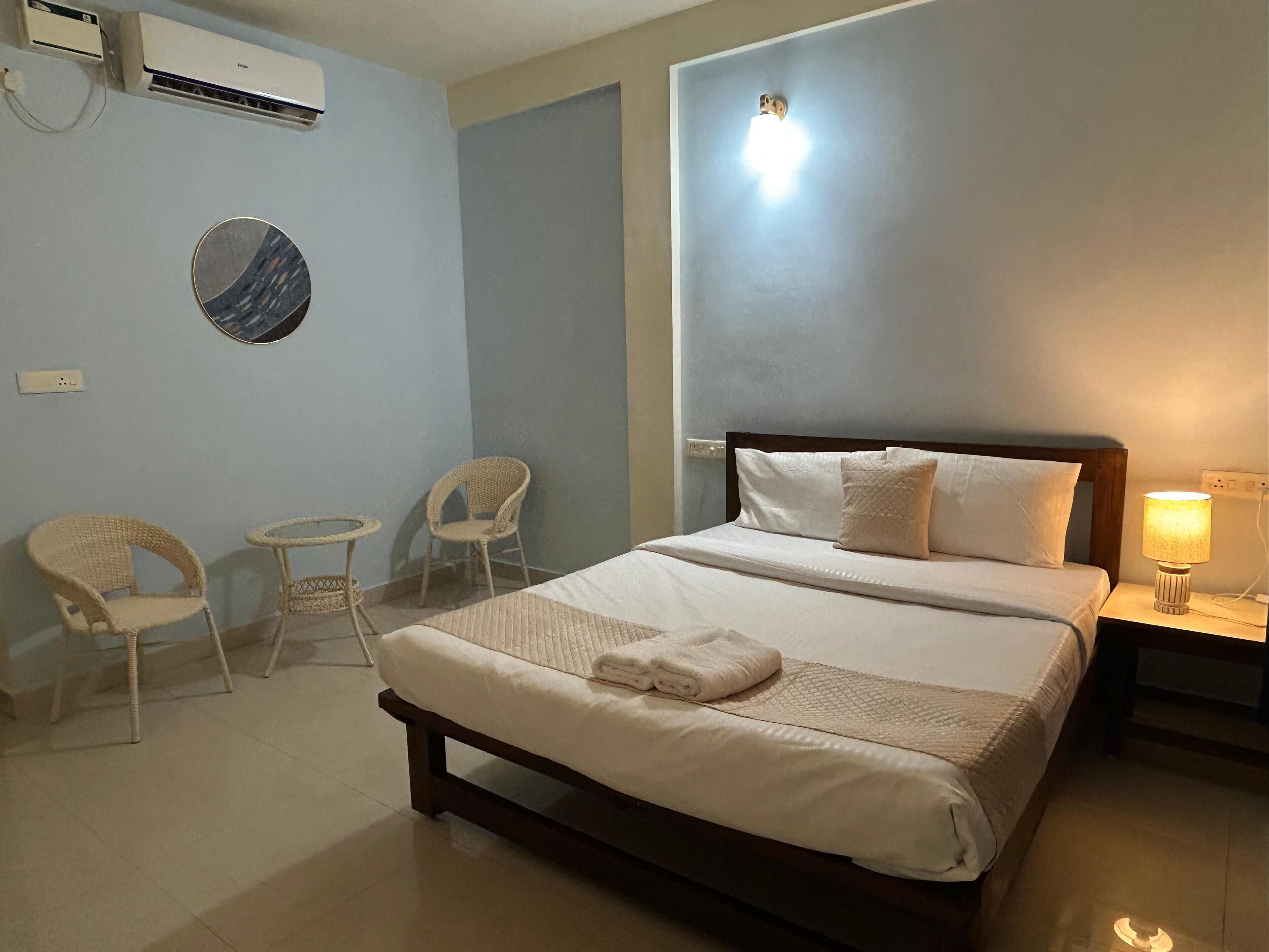Habitación de hotel con cama, dos sillones de mimbre, una mesita redonda, aire acondicionado de pared y una lámpara de noche.