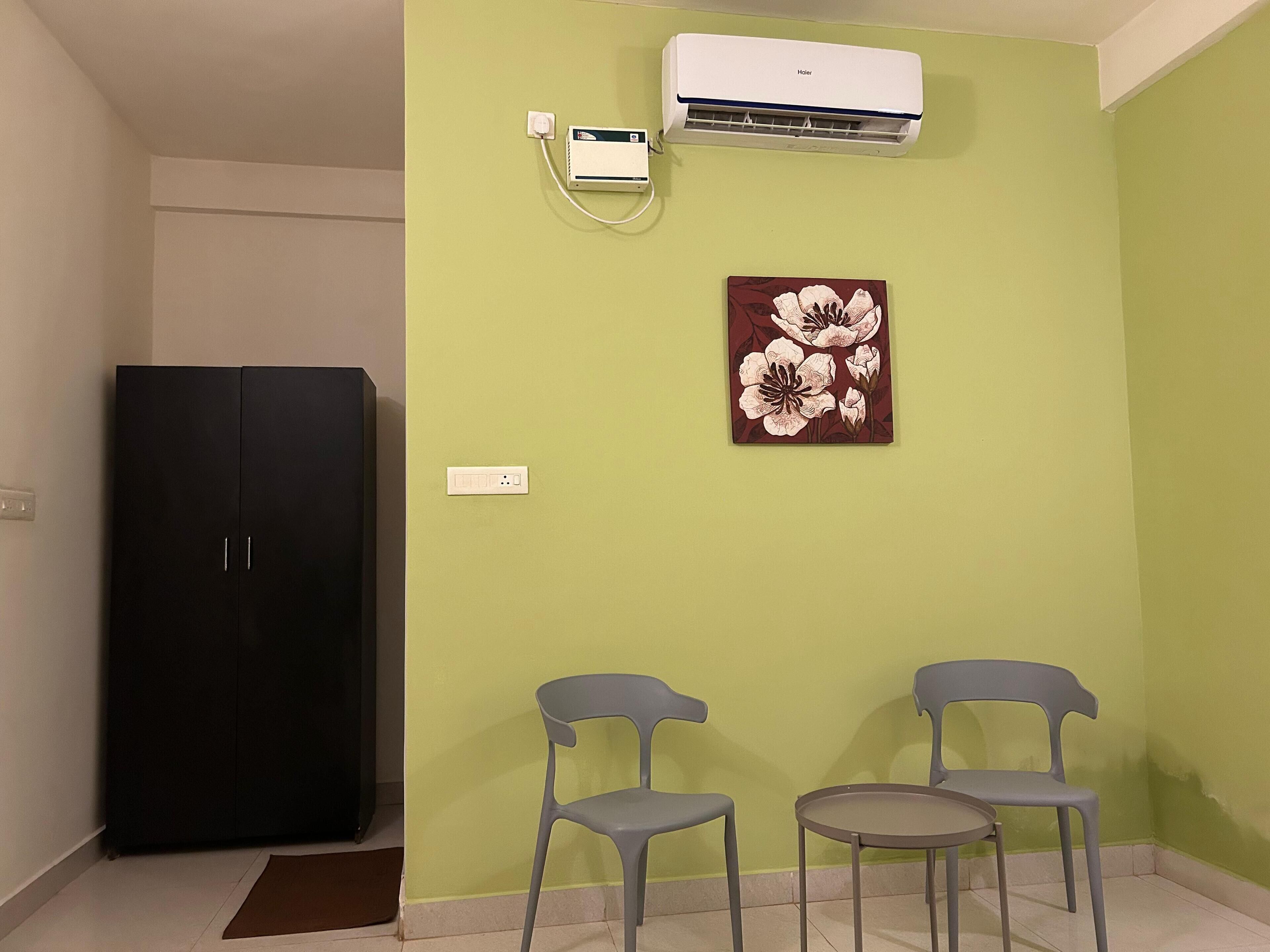 A room with a black wardrobe, two grey chairs, a small round table, a wall-mounted air conditioner, and a floral painting.