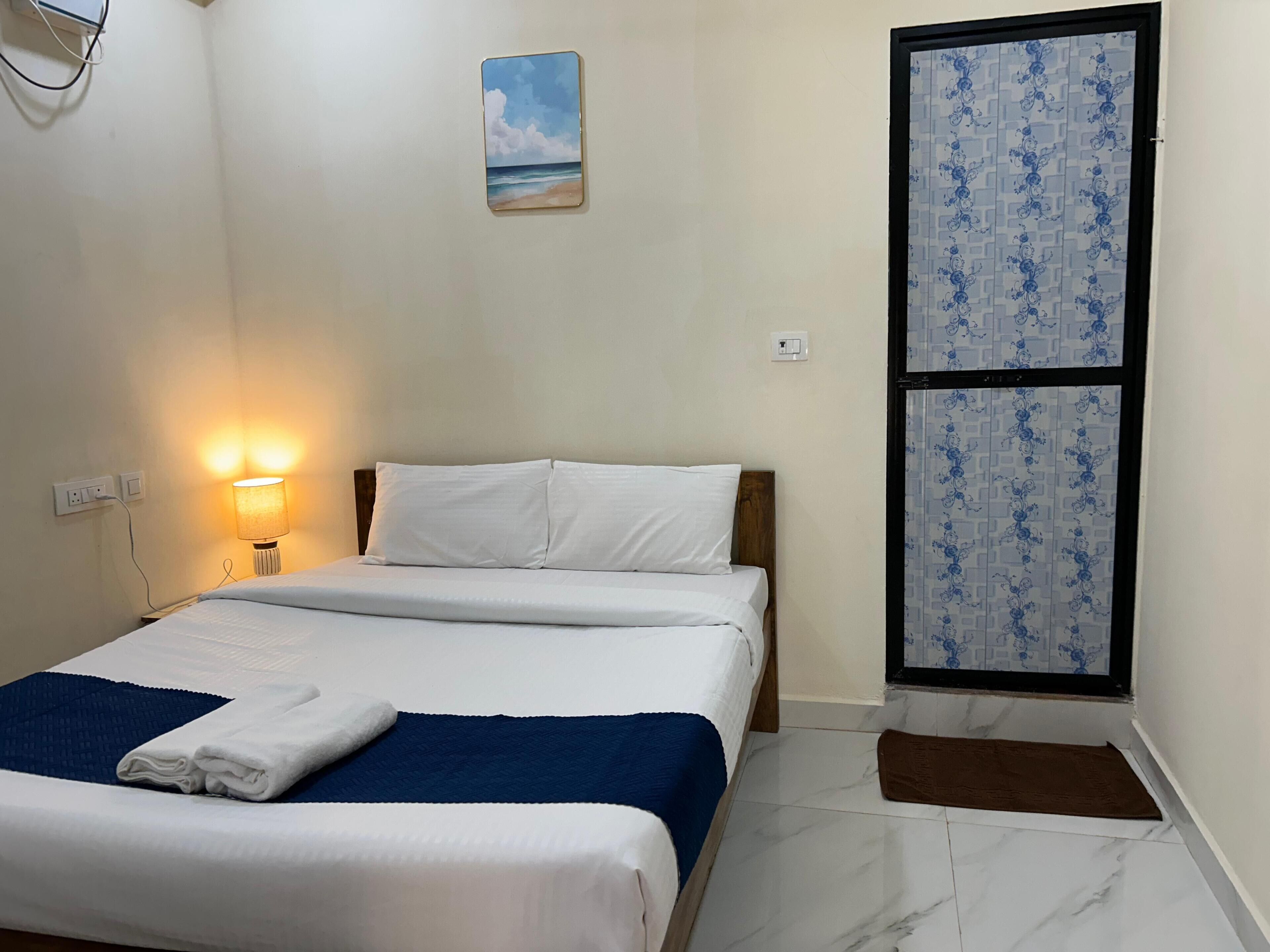 A neatly made bed with white and blue bedding, a bedside lamp, and a painting of a beach scene on the wall.