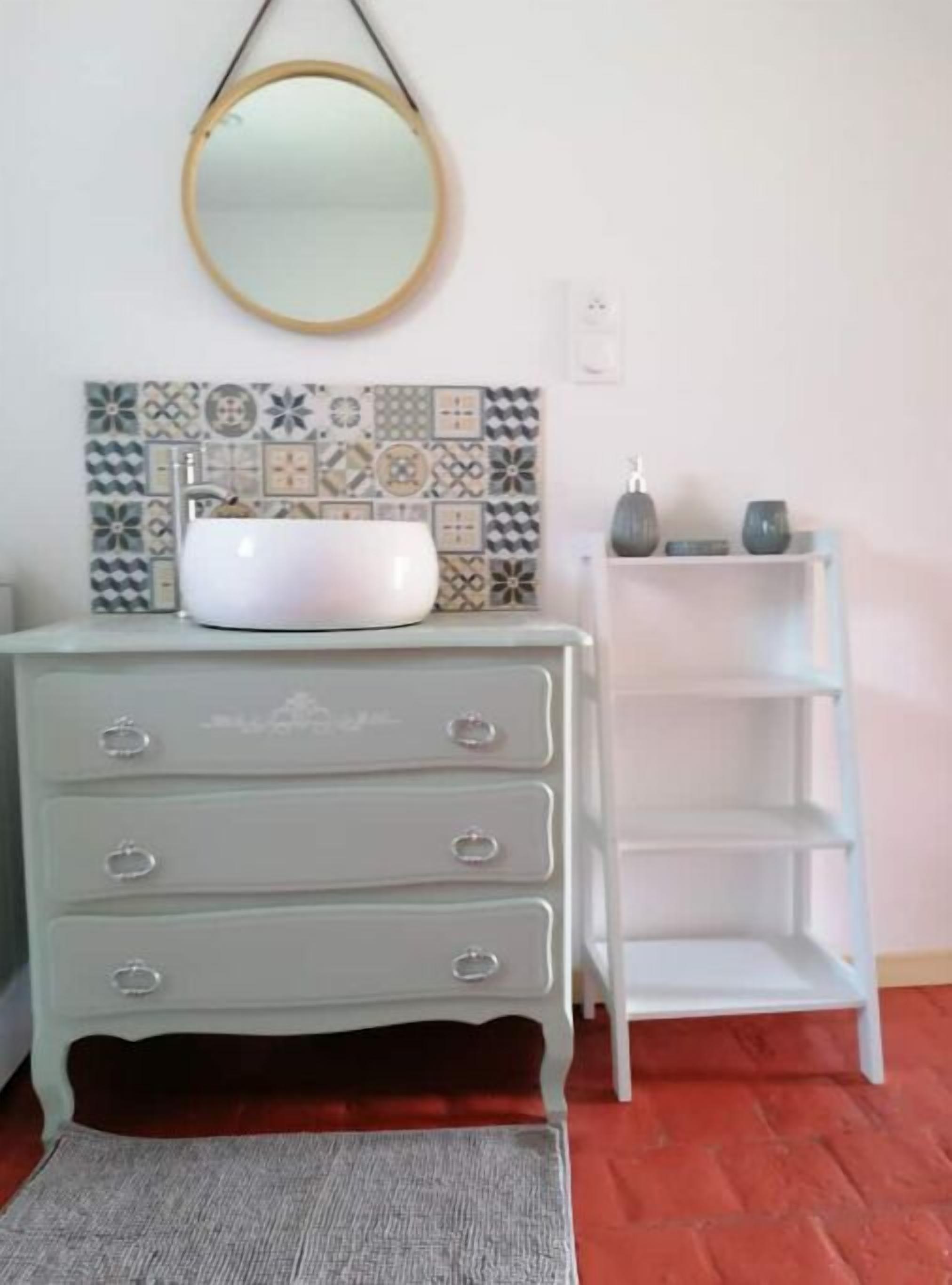 A bathroom with a white sink, a mirror, a patterned tile backsplash, a light blue vanity with drawers, a white shelf, and a gray rug on a red floor.