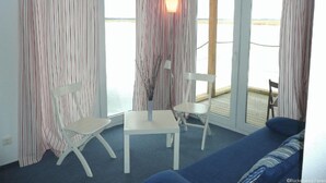 A room with a blue sofa, two white chairs, a small table, and a view of the sea through large windows with striped curtains.
