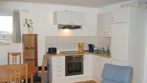 A compact kitchen with a stove, oven, sink, and white cabinets. There is a dining table with chairs and a blue chair. - Floating Houses - Komfortable Schwimmende Ferienhäuser im Krösliner Yachthafen (Kröslin)