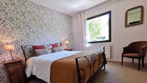 Une chambre à coucher comprenant un lit, deux tables de chevet, une chaise, une fenêtre et un mur orné d’un motif floral.