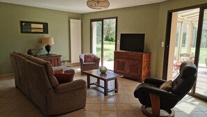 Un salon comprenant un canapé, un fauteuil, une table basse et un téléviseur posé sur un meuble en bois. Une fenêtre offre une vue sur la verdure à l’extérieur.