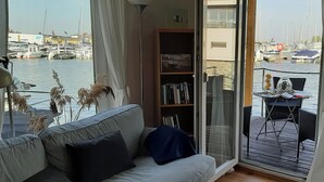 Un salon avec un canapé, une bibliothèque et une vue sur une marina, visible à travers une porte vitrée.