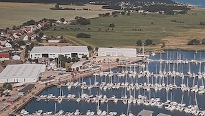 Un port de plaisance où sont amarrés de nombreux bateaux, un grand bâtiment doté d’un toit plat et un paysage environnant composé de champs et de collines.