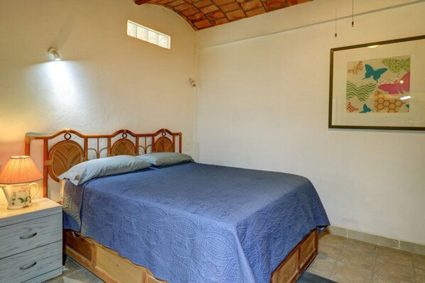 A bedroom with a wooden bed, a nightstand with a lamp, and a framed butterfly artwork on the wall. - Casa las Mariposas Bungalow 6 (Los Ayala)