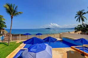 A pool area with blue umbrellas, a clear view of the ocean, and palm trees. - Casa las Mariposas Bungalow 6 (Los Ayala)