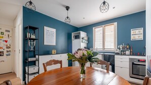 Un comedor con una mesa de madera, sillas, un florero con flores, un refrigerador blanco, un microondas, una estufa y una ventana con persianas.