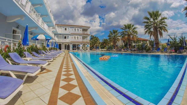 A swimming pool with lounge chairs and umbrellas, a building with balconies, and palm trees in the background. - Laganas Holidays Hotel (Zakynthos)