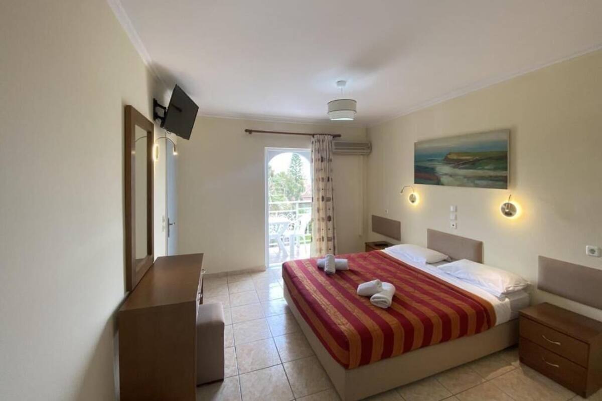 Quarto de hotel com cama, escrivaninha, cadeira, televisão, quadro e janela com cortinas.