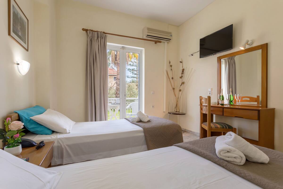 Quarto de hotel com duas camas, uma escrivaninha com cadeira, televisão e janela com cortinas.