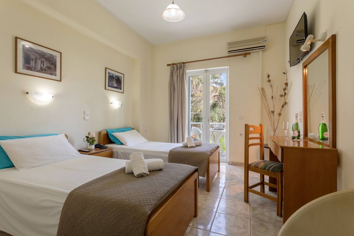 Quarto de hotel com duas camas, uma escrivaninha, uma cadeira, um espelho e uma varanda com vista.