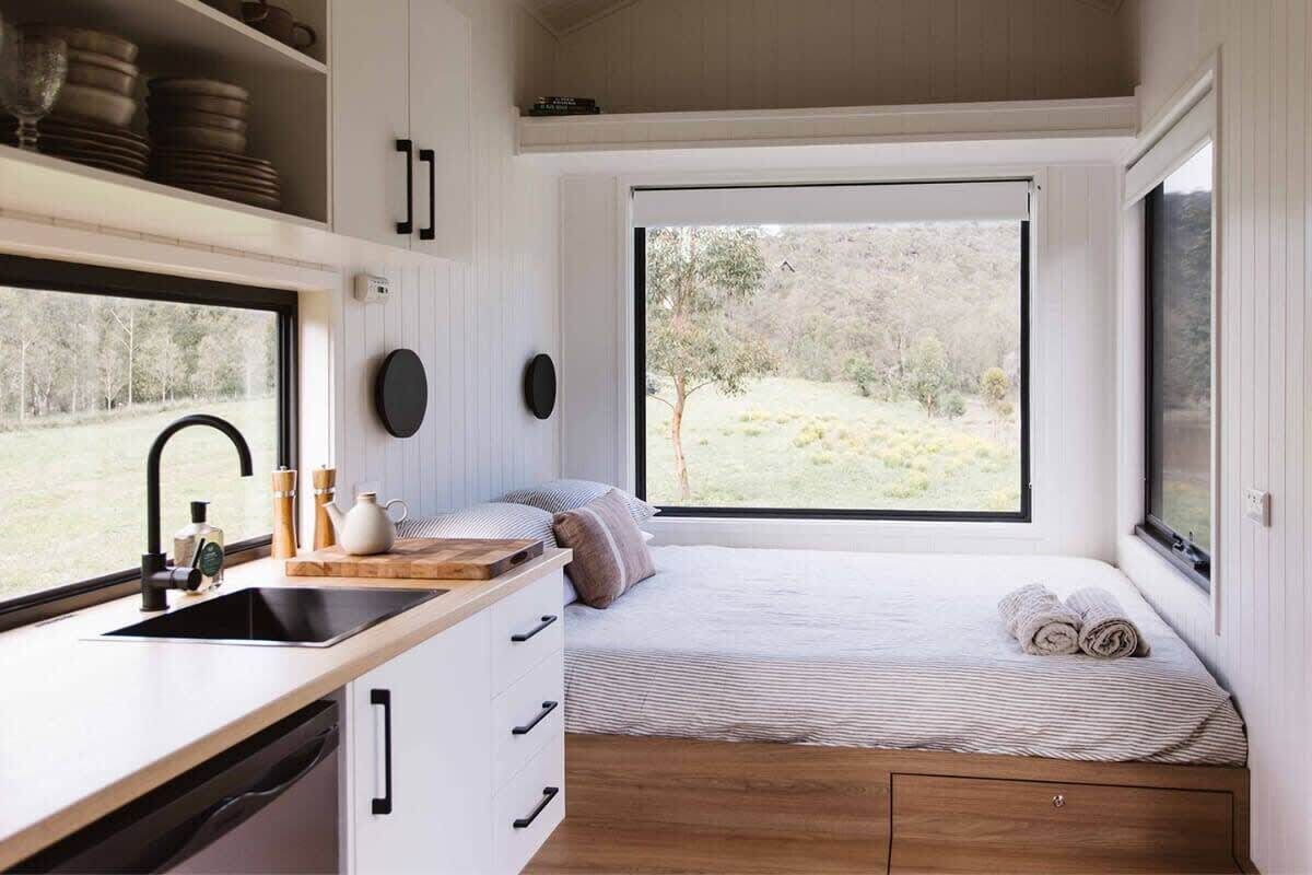 A compact bedroom with a window, a bed, a bedside table, and a built-in kitchen area.