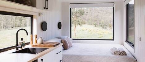 A compact bedroom with a window, a bed, a bedside table, and a built-in kitchen area.