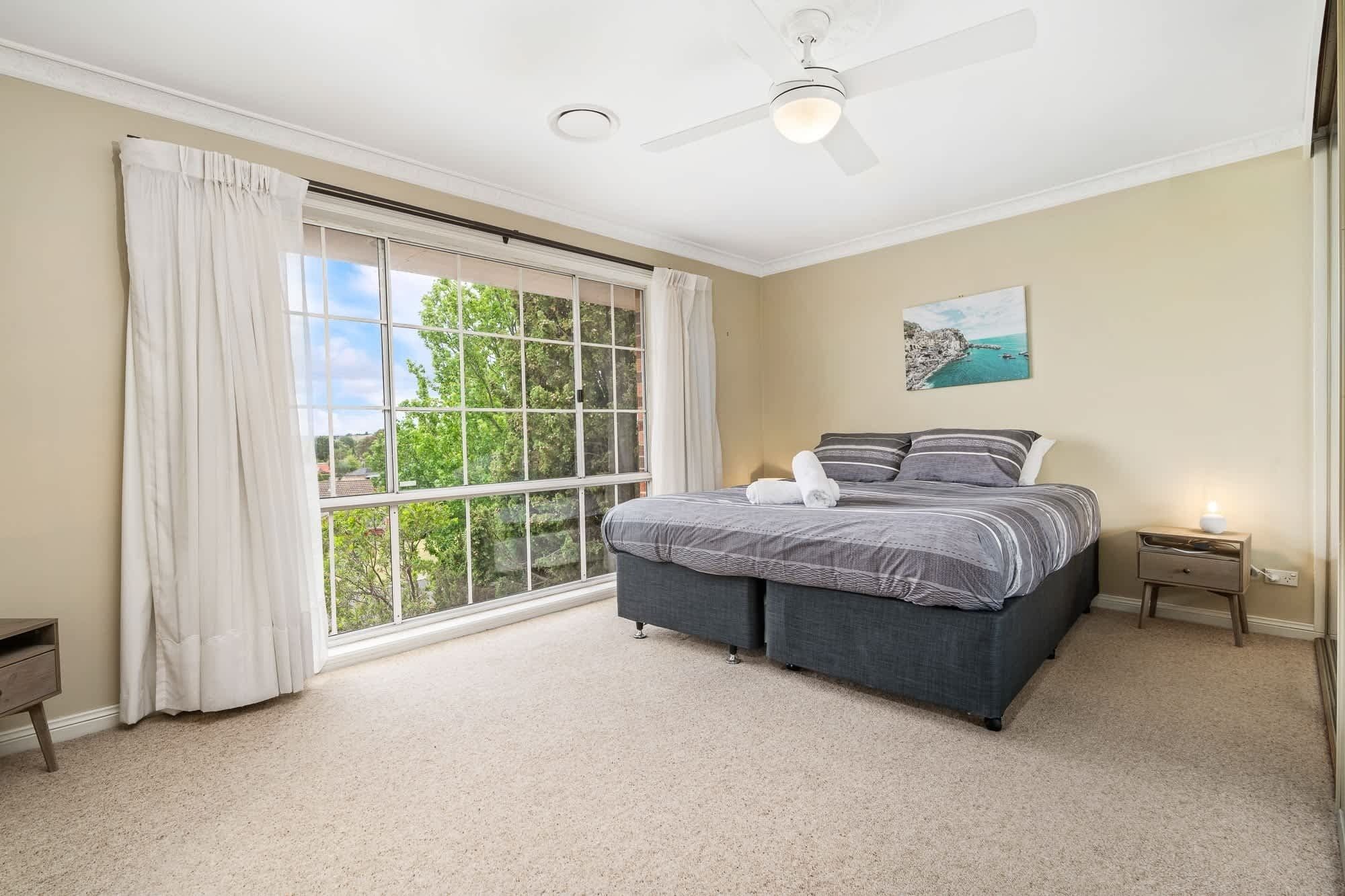 A bedroom with a large window, a bed, a ceiling fan, and a nightstand.