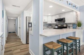 A modern kitchen with white cabinets, a stainless steel refrigerator, and a microwave. There are three stools with woven seats at the counter.