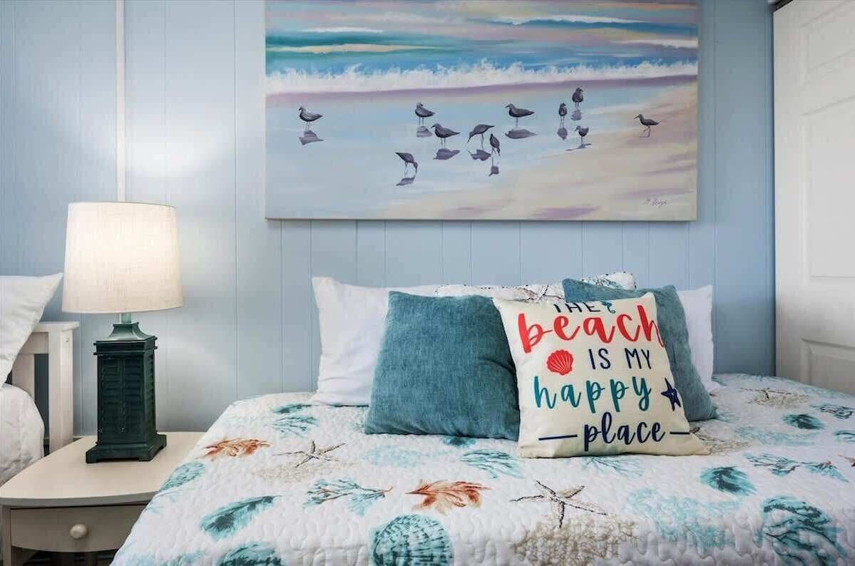 Quarto com colcha decorada com tema praiano, pintura de pássaros na parede e almofada com frase sobre a praia.