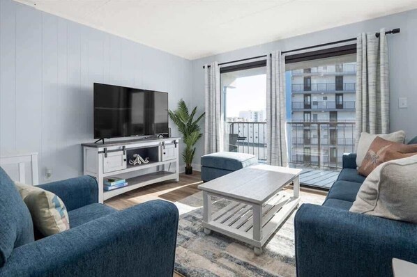 A modern living room with a flat-screen TV, blue sofas, a coffee table, and a large window with a balcony view. - Wave Watcher's Condo - Steps to the beach! (Ocean City)